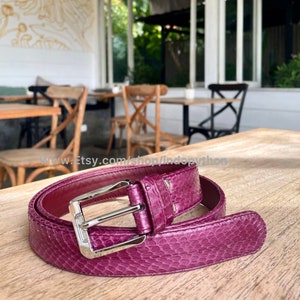 Peut inclure: Une ceinture en cuir de serpent violet avec une boucle argentée. La ceinture est posée sur une table en bois.
