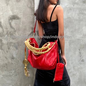 Puede incluir: Un bolso de mano de cuero rojo con una correa de cadena dorada y un borla de moneda de oro. El bolso lo lleva una persona que lleva un vestido negro.