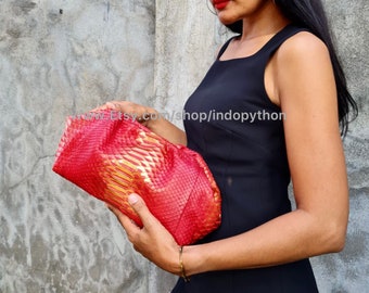 May include: A red and gold python skin clutch purse. The purse is folded in half and held by a person's hand. The purse has a textured, scaly surface.