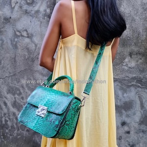 May include: A green snakeskin handbag with a silver clasp and a long strap. The bag is worn by a person wearing a yellow dress.
