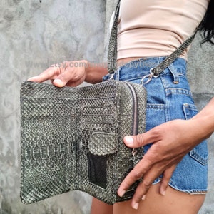 Pode incluir: Uma carteira de pele de cobra cinza com fecho de correr e uma alça comprida. A carteira está aberta e mostra vários slots para cartões e um bolso com fecho de correr.