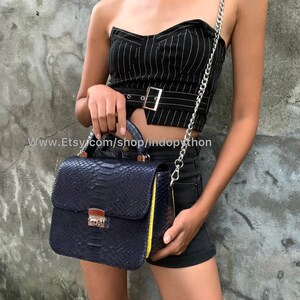 May include: A dark blue snakeskin handbag with a yellow trim and a silver chain strap. The bag has a top handle and a flap closure with a silver lock.