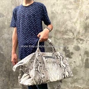 Puede incluir: Una bolsa de viaje de cuero de serpiente blanca y gris con cierre de cremallera y una correa para el hombro. La bolsa está siendo sostenida por una persona que lleva una camisa con estampado azul y blanco.