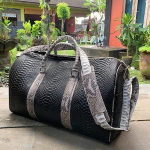 Puede incluir: Una bolsa de viaje de cuero negro con un patrón de piel de serpiente y correas de piel de serpiente marrón claro. La bolsa tiene un asa superior y una correa para el hombro.