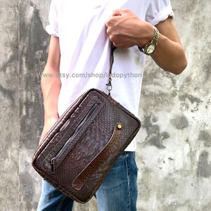 May include: A brown leather clutch bag with a snakeskin pattern and a long strap. The bag has a zipper closure and a small pocket on the front. The bag is made of genuine leather and has a unique design.