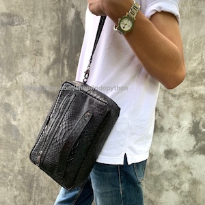 Può includere: Una pochette nera in pelle di serpente con una cerniera argentata e una lunga tracolla. La borsa è indossata sulla spalla di una persona che indossa una maglietta bianca e jeans blu. www.etsy.com/shop/indopython