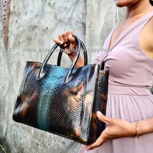 Może przedstawiać: Czarna, zielona i brązowa torba typu tote ze skóry węża z górnym uchwytem. Torba ma błyszczące, tęczowe wykończenie.