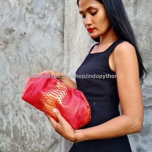 Könnte beinhalten: Eine rote Clutch aus Pythonleder mit goldenen Akzenten. Die Clutch ist in der Mitte gefaltet und wird von einer Hand gehalten. Die Clutch hat eine strukturierte, schuppige Oberfläche.