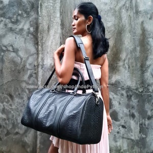 Puede incluir: Una bolsa de viaje de cuero negro con un patrón de piel de serpiente. La bolsa tiene una correa de hombro ajustable larga y un asa superior. La bolsa está siendo sostenida por una persona que lleva un vestido rosa.