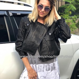 Może przedstawiać: Czarna skórzana kurtka ze wzorem imitującym skórę węża. Kurtka ma zamek błyskawiczny i kołnierz. Modelka nosi kurtkę na białym t-shircie z nadrukiem graficznym. Tekst na t-shircie to "GION".