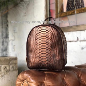 Może przedstawiać: Brązowy skórzany plecak ze wzorem skóry węża. Plecak ma zamek błyskawiczny i górny uchwyt.