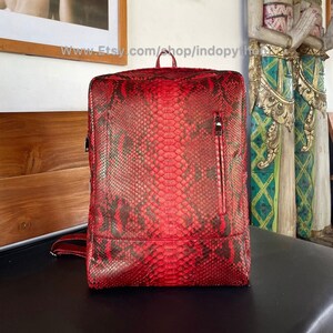 Könnte beinhalten: Ein roter Schlangenleder-Rucksack mit einer Reißverschlusstasche auf der Vorderseite. Der Rucksack ist aus echtem Pythonleder gefertigt und hat ein einzigartiges, strukturiertes Muster.