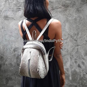 May include: A white python leather backpack with a zipper closure. The backpack has a unique, textured design and is worn by a person.