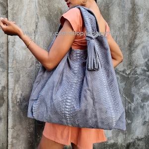 Puede incluir: Un bolso tote grande de piel de serpiente gris con un largo borlón. El bolso tiene una forma holgada y sin estructura y está hecho de piel suave y flexible.