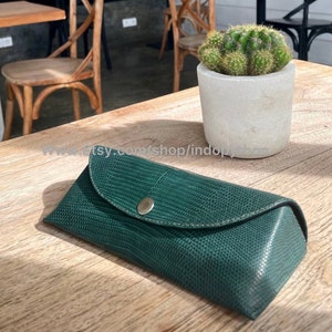 Könnte beinhalten: Ein dunkelgrünes, strukturiertes Brillenetui mit Druckknopfverschluss. Das Etui besteht aus einem Material, das Reptilienhaut imitiert. Das Etui ist geschlossen und liegt auf einer Holzoberfläche. Ein kleiner Kaktus in einem Betonbehälter befindet sich im Hintergrund.