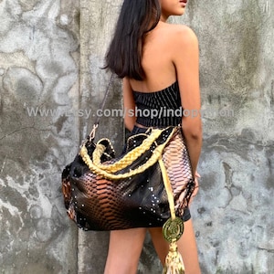 Può includere: Una borsa a mano in pelle di serpente nera e marrone con dettagli dorati e un pendente a nappa dorato. La borsa ha una tracolla intrecciata e viene indossata sulla spalla.