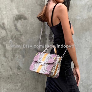 Op de afbeelding: Een klein, roze, geel en wit snakeskin handtasje met een zilveren kettingband. De tas heeft een bovenste handvat en een klep sluiting.