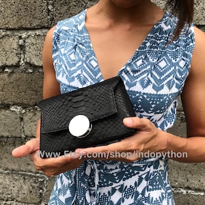 May include: A black python skin clutch with a silver circular clasp. The clutch is held in the hands of a person wearing a blue and white patterned dress.