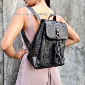 Może przedstawiać: Czarny skórzany plecak ze wzorem skóry węża i frędzlem. Plecak ma klapkę zamykaną na metalową klamrę.
