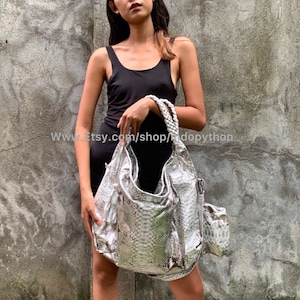 Op de afbeelding: Een zilveren tote bag van pythonleer met een franje detail. De tas heeft een bovenste handvat en een lange band. De tas wordt vastgehouden door een persoon die een zwart tanktop draagt.