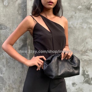 May include: A black snakeskin clutch purse with a gathered, ruched design. The purse is held by a person wearing a black one-shoulder top and black pants.