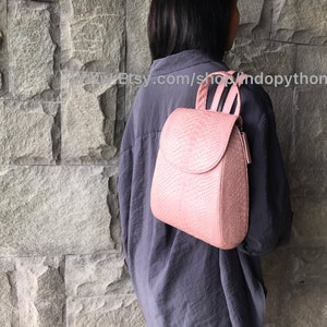 Może przedstawiać: Różowy plecak skórzany o unikalnym, zakrzywionym kształcie i fakturowanym wzorze skóry węża. Plecak ma górny uchwyt i regulowane paski.