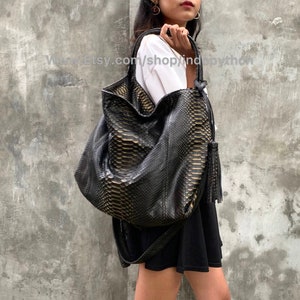 Op de afbeelding: Een grote zwarte en gouden slangleren tote bag met een kwast. De tas heeft een slonzige, ongestructureerde vorm en wordt over de schouder gedragen.