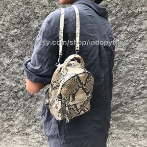 Könnte beinhalten: Ein kleiner, beige-schwarzer Schlangenleder-Rucksack mit Reißverschluss. Der Rucksack hat einen oberen Griff und verstellbare Träger.