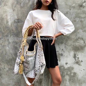 May include: A white cropped sweater with long sleeves and a white and grey python skin handbag with a gold braided strap. The handbag has a tassel charm.