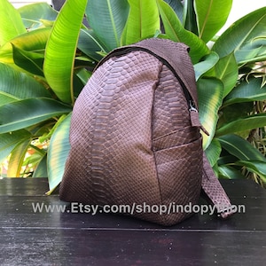 Könnte beinhalten: Brauner Rucksack mit Schlangenmuster. Die Tasche hat einen Reißverschluss und verstellbare Träger. Der Rucksack besteht aus einem strukturierten Material, vor dem Hintergrund grüner Blätter.