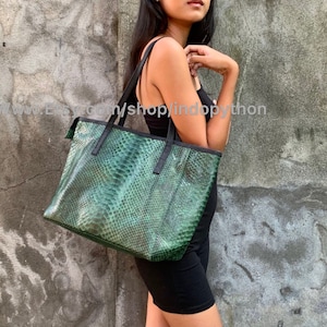 May include: A green snakeskin tote bag with black leather handles and trim. The bag is being held by a person wearing a black dress.