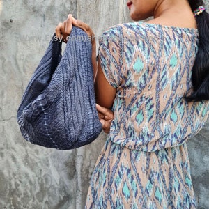 Könnte beinhalten: Eine dunkelblaue, strukturierte Handtasche mit geschwungener Form. Die Tasche scheint aus einem Material zu bestehen, das Reptilienhaut ähnelt. Der Hintergrund ist eine schlichte, graue Wand. Die Tasche wird von einer Person gehalten, die ein gemustertes Kleid trägt.