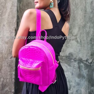 Może przedstawiać: Różowy plecak ze skóry węża z złotym zamkiem błyskawicznym. Plecak jest mały i ma zaokrąglony kształt. Plecak jest noszony przez osobę.