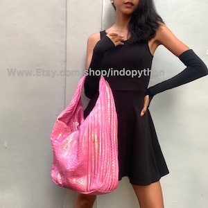 Puede incluir: Un bolso tote de piel de pitón rosa con un acabado brillante. El bolso es grande y tiene una forma holgada. El bolso lo lleva una persona que lleva un vestido negro y guantes largos negros.