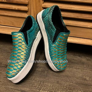 May include: A pair of teal and gold slip-on sneakers with a scale-like pattern. The shoes have white soles and black interior lining. The Etsy shop URL is visible on the side of the shoe.