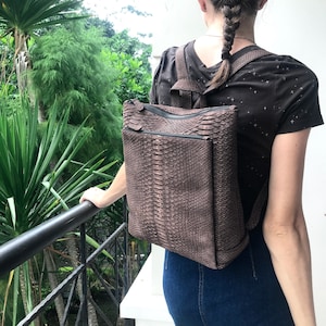 Könnte beinhalten: Ein brauner Schlangenleder-Rucksack mit Reißverschluss. Der Rucksack wird von einer Person mit langen braunen Haaren getragen.