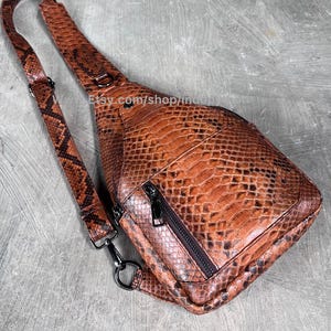 Könnte beinhalten: Eine braune und schwarze Sling-Bag mit Schlangenmuster, mit einem schwarzen Reißverschluss und Riemen. Die Tasche hat einen einzelnen Riemen und eine kleine Reißverschlusstasche. Die Tasche besteht aus einem strukturierten Material und hat ein schlankes Design.