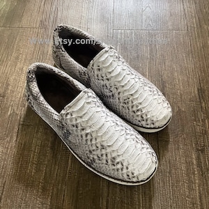 Op de afbeelding: Een paar witte slangenleren instappers met een bruine leren voering. De schoenen hebben een witte rubberen zool.
