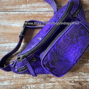 May include: A purple snakeskin fanny pack with black zipper accents and a black adjustable strap. The bag has a small front pocket with a zipper closure. The bag is on a wooden surface.