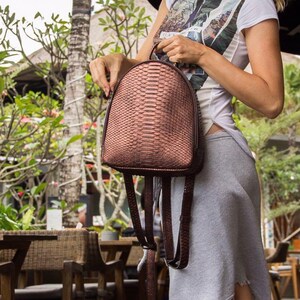 Könnte beinhalten: Ein brauner Schlangenleder-Rucksack mit Reißverschluss und verstellbaren Trägern. Der Rucksack wird von einer Person in einem zwanglosen Umfeld getragen.