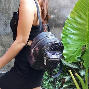 Könnte beinhalten: Ein schwarzer Lederrucksack mit Schlangenhautmuster. Der Rucksack hat eine runde Form und einen Reißverschluss.