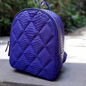 Może przedstawiać: Fioletowy plecak ze skóry węża z pikowanym wzorem. Plecak ma zamek błyskawiczny i regulowane paski.