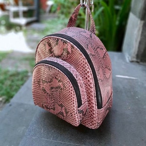 Może przedstawiać: Mały, różowo-brązowy plecak z wzorem skóry węża. Plecak ma zaokrąglony kształt z przednią kieszenią i czarnymi zamkami błyskawicznymi. Ma górny uchwyt i jest wystawiony na zewnątrz.