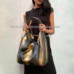 Op de afbeelding: Een zwarte en gouden slangleren hobo-tas met een geplisseerd ontwerp. De tas wordt vastgehouden door een persoon die een zwarte mouwloze jurk draagt.