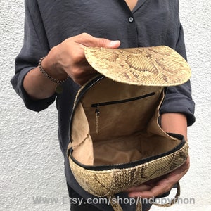 Puede incluir: Una mochila de piel de serpiente de color beige con cierre de cremallera y un peque&ntilde;o bolsillo en el interior. La mochila est&aacute; abierta y el interior es visible.