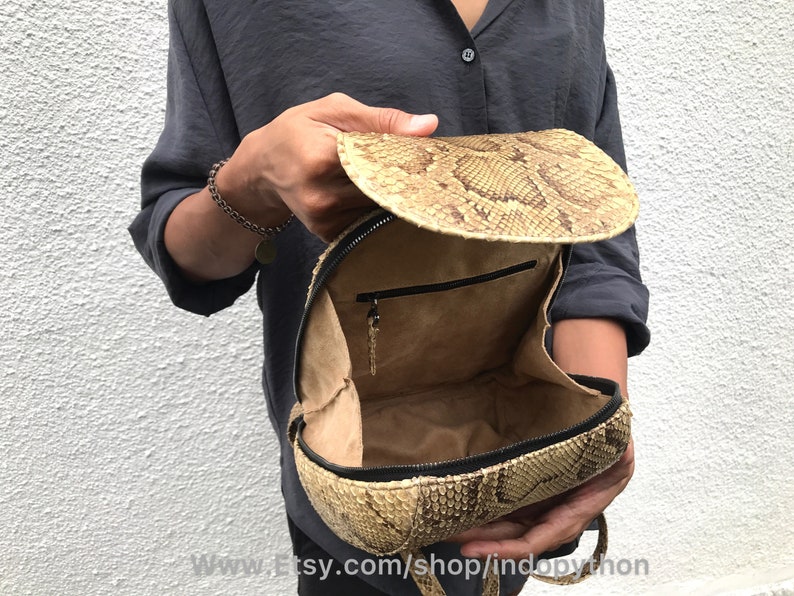 Puede incluir: Una mochila de piel de serpiente de color beige con cierre de cremallera y un peque&ntilde;o bolsillo en el interior. La mochila est&aacute; abierta y el interior es visible.