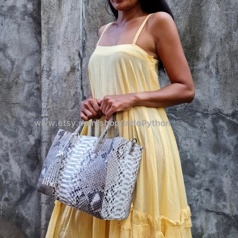 May include: A woman in a yellow dress holds a light brown snakeskin tote bag with a tassel. The bag has a top handle and a shoulder strap. The text "www.etsy.com/shop/IndoPython" is visible in the image.