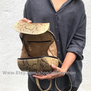 Puede incluir: Una mochila de piel de pit&oacute;n beige con un bolsillo con cremallera en el interior. La bolsa est&aacute; abierta y el interior es visible. La bolsa tiene un asa corta y una correa larga.