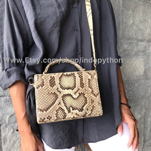 May include: A snakeskin-patterned handbag in tan, brown, and cream. The bag has a rectangular shape, a matching strap, and a small handle. The text "www.Etsy.com/shop/indopython" is visible on the image.