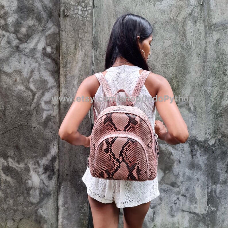 K&ouml;nnte beinhalten: Ein rosafarbener Rucksack aus Pythonleder mit Rei&szlig;verschluss und verstellbaren Tr&auml;gern. Der Rucksack wird von einer Person getragen, die vor einer Betonwand steht.